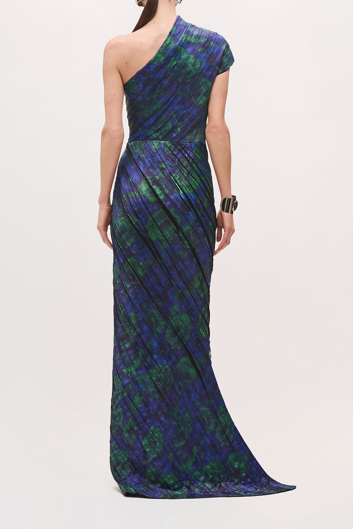 Back view of a model wearing the Karina Dress in Blue Floral with one sleeve, diagonal pleats throughout the dress, and a floor-length skirt.
