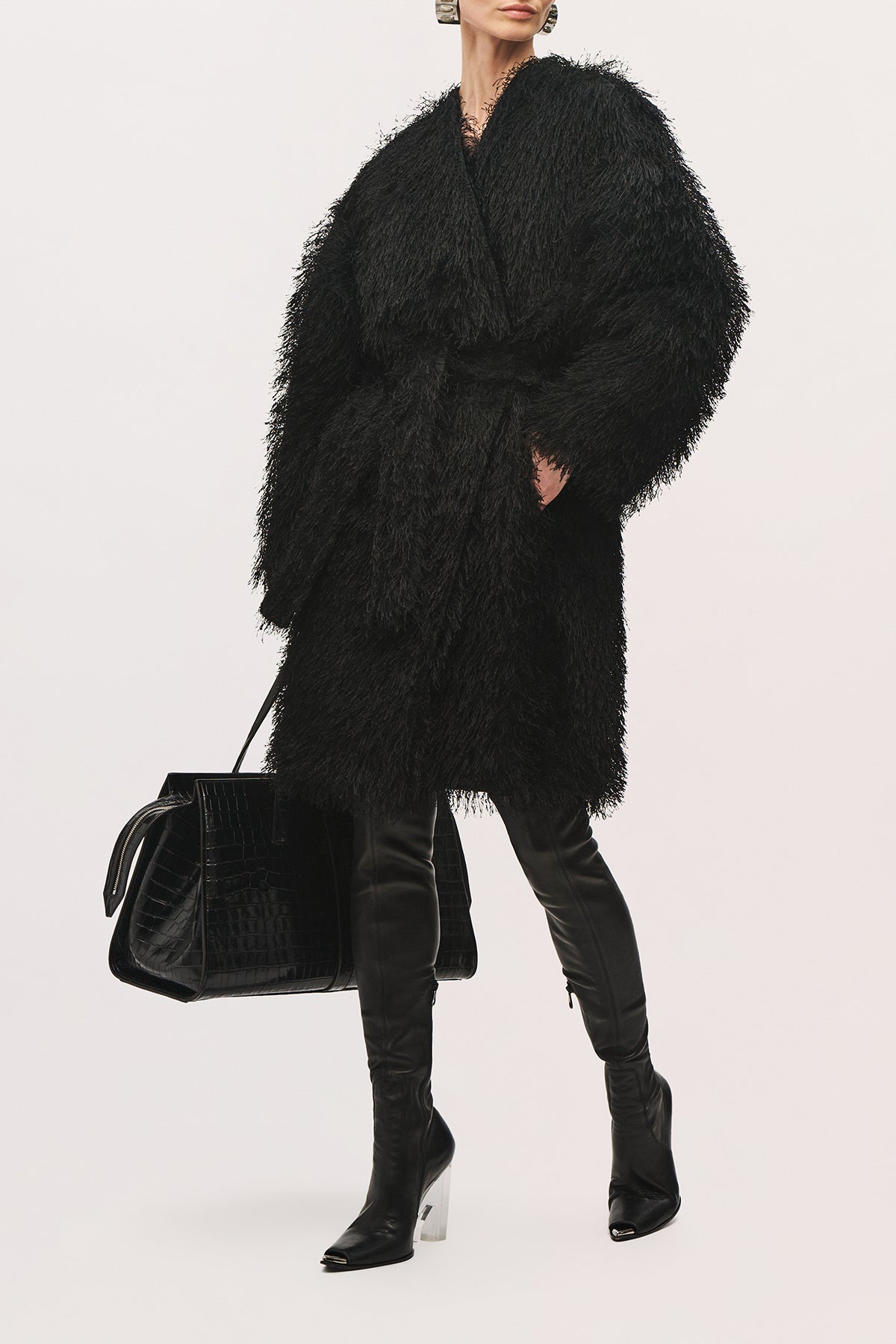 Angled front view of a model wearing the Sabrina Coat in Black, showing its voluminous wrap silhouette, textured fringe surface, wide sleeves, self-tie waist, and knee-length hem.
