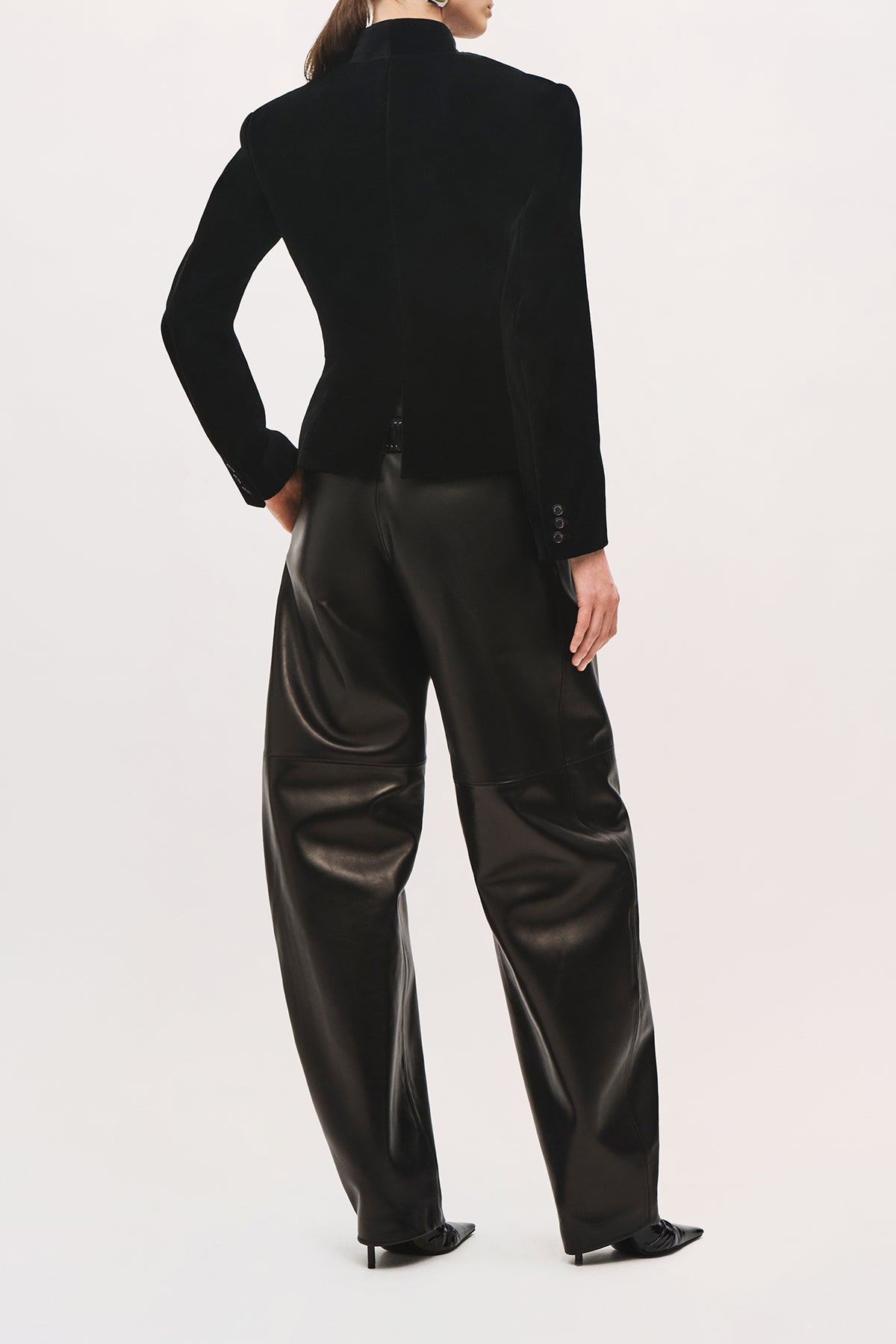 Back view of a model wearing the Alora Jacket in Black, showing its fitted velvet silhouette, structured shoulders, high collar, shaped waist seams, center-back vent, and buttoned cuffs.