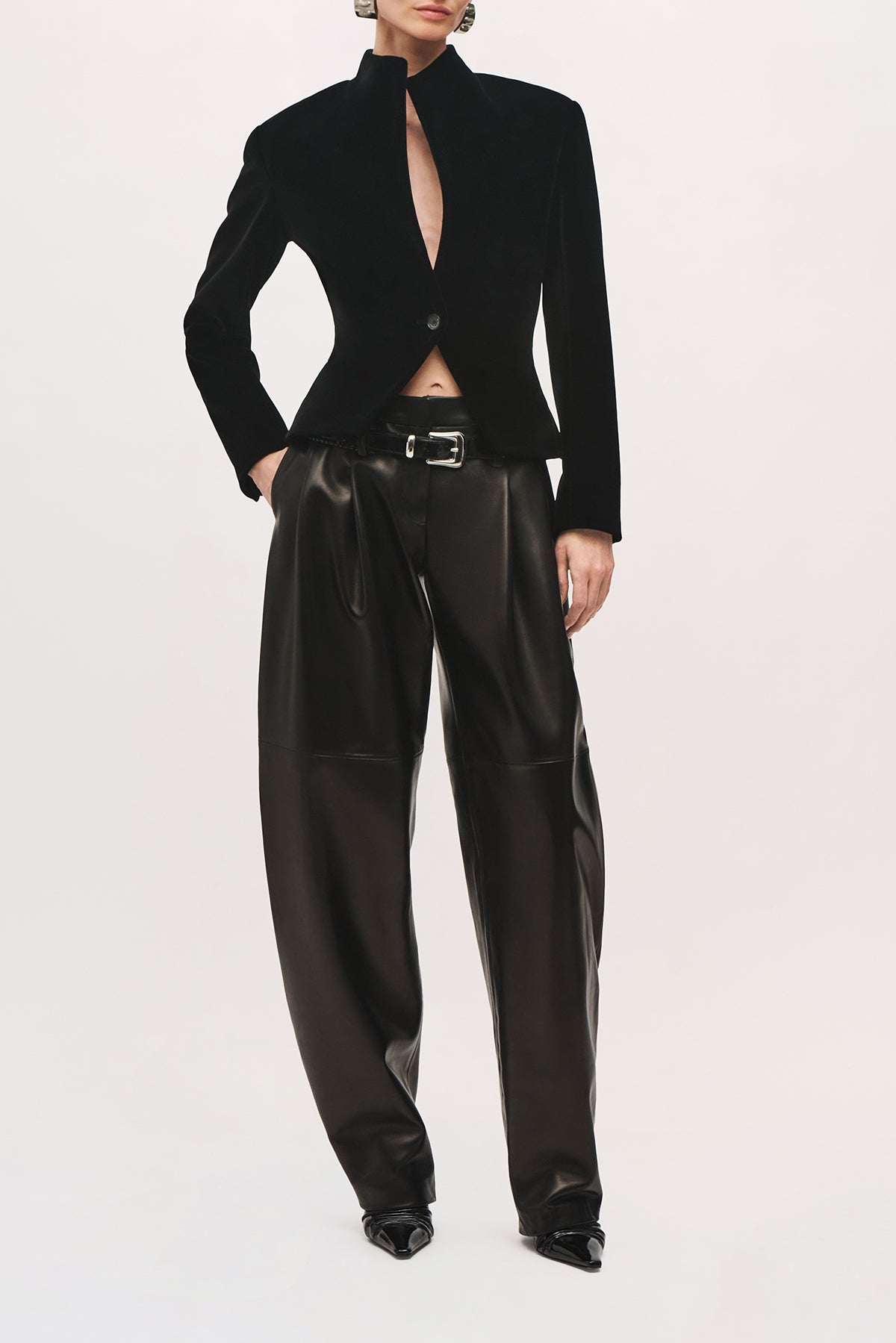 Front view of a model wearing the Alora Jacket in Black, a fitted velvet jacket with structured shoulders, a high stand collar, deep front keyhole opening, single-button closure, and curved hem.