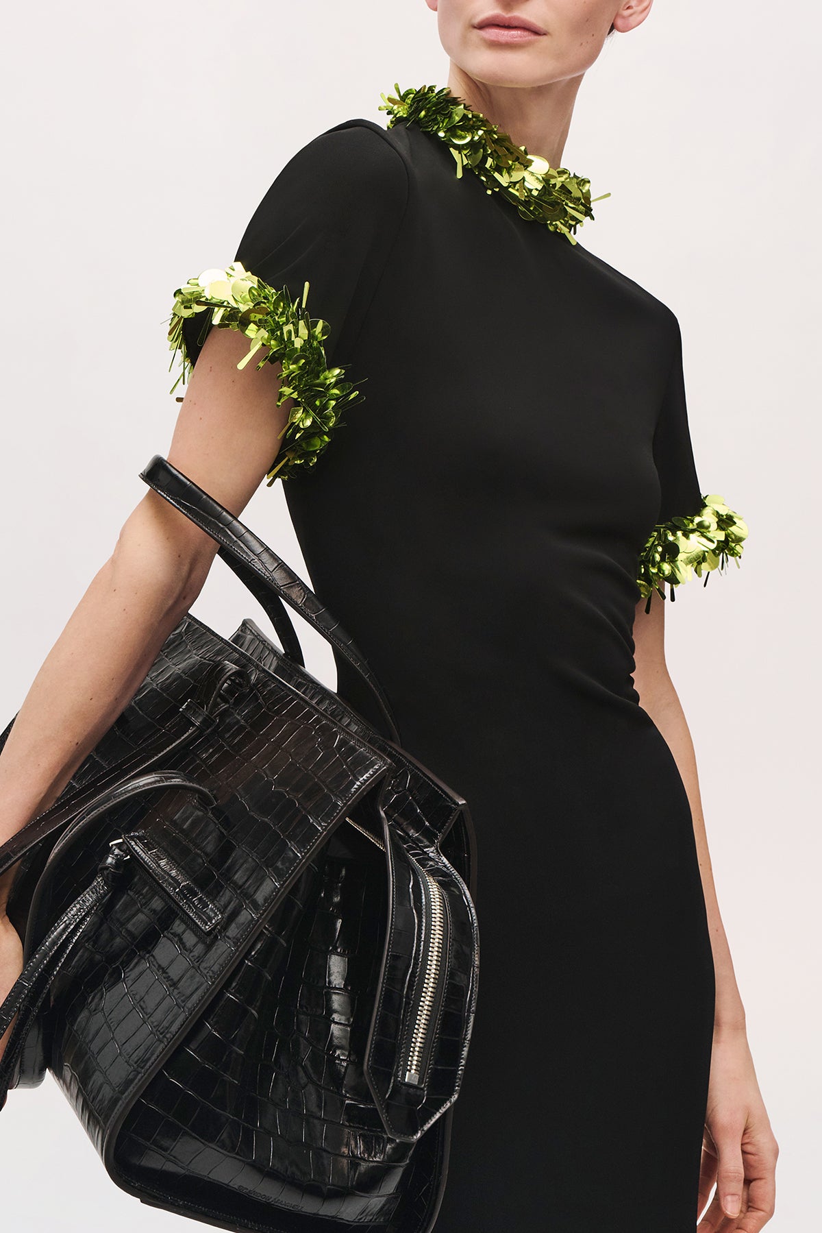 Close-up angled front view of a model holding a black purse and wearing the Bex Gown in Black/Green Glo, highlighting the high neckline, short sleeves, and green sequin trim.