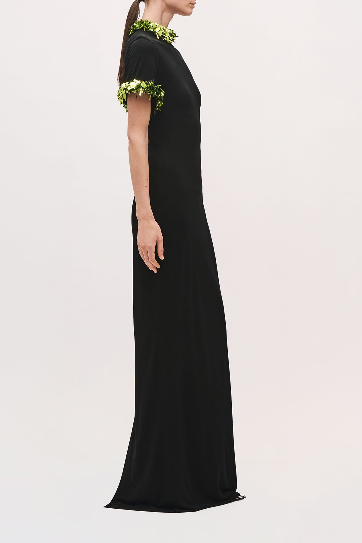 Side view of a model wearing the Bex Gown in Black/Green Glo, showing its slim floor-length silhouette and green sequin-trimmed sleeves.