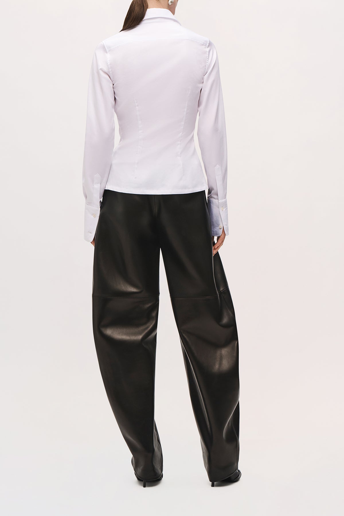 Back view of a model wearing the Ceci Button Down in White, showing its fitted silhouette, pointed collar, back shaping seams, long sleeves, and extended buttoned cuffs.