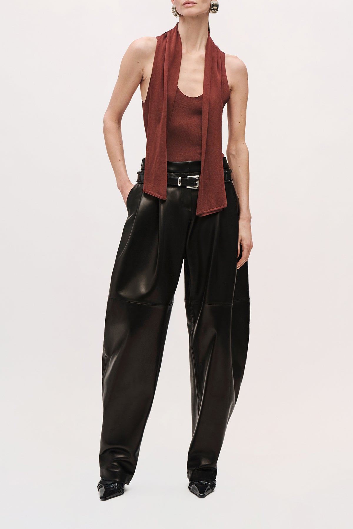 Front view of a model wearing the Dean pants in black nappa leather with a low-rise waist and relaxed, curved-leg silhouette, and a silver belt buckle. 