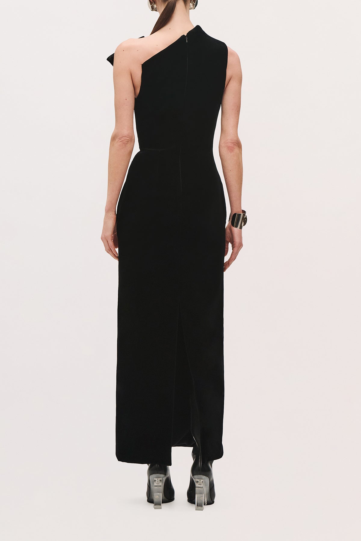 Back view of a model wearing the Farren Dress in Black, showing its sleeveless silhouette, with an asymmetrical one shoulder detail, and long straight fit. 
