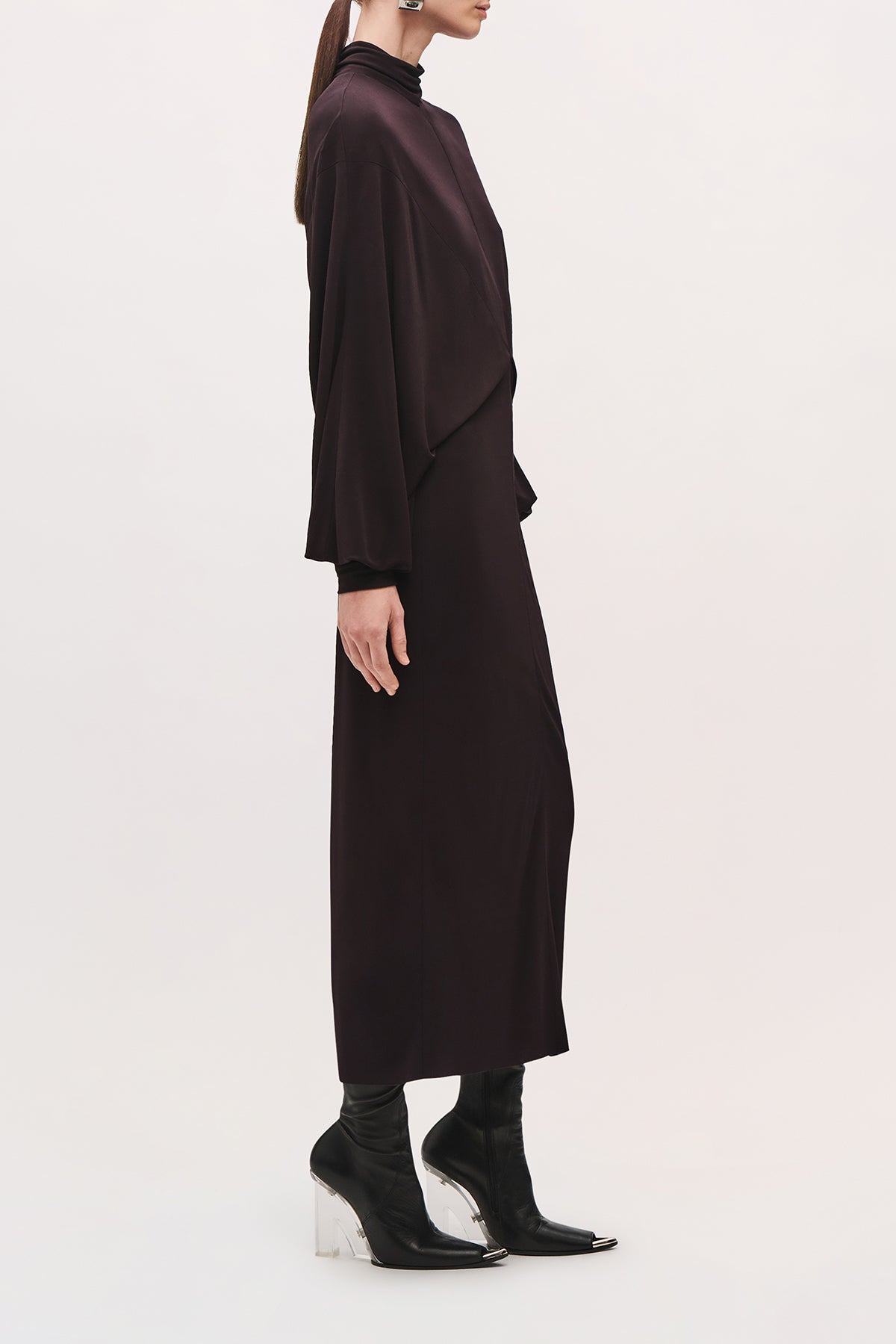 Side view of a model wearing the Helena Dress in Ganache, showing its high neckline, draped long-sleeve bodice, and straight midi-length skirt.