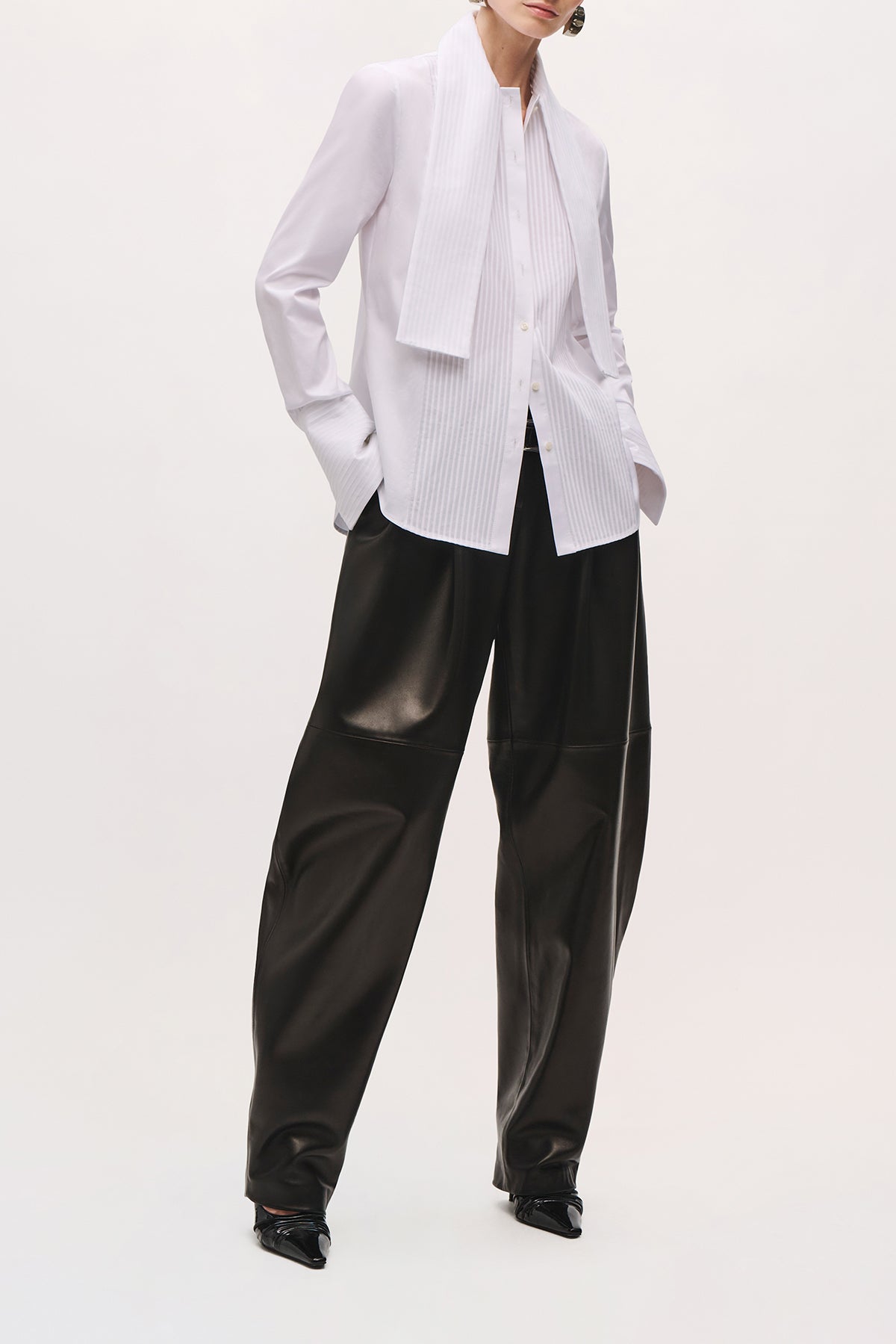 Front view of a model wearing the Inez Shirt in White with buttons down the front, vertical pleats, and an attached scarf. 