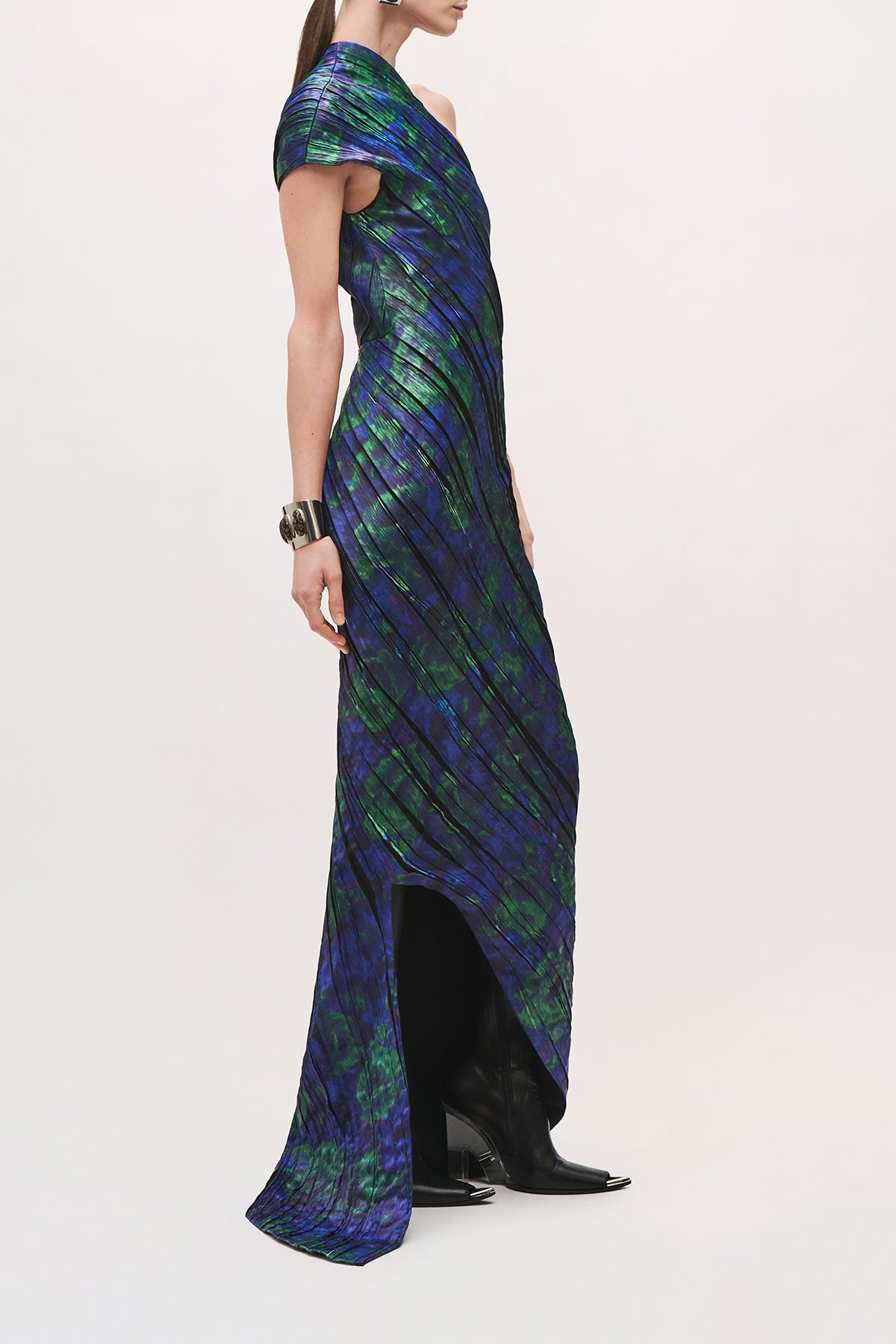 Side product view of a model wearing the Karina Dress in Blue Floral, with one draped cap sleeve, fitted silhouette, side slit, and asymmetric floor-length skirt. 