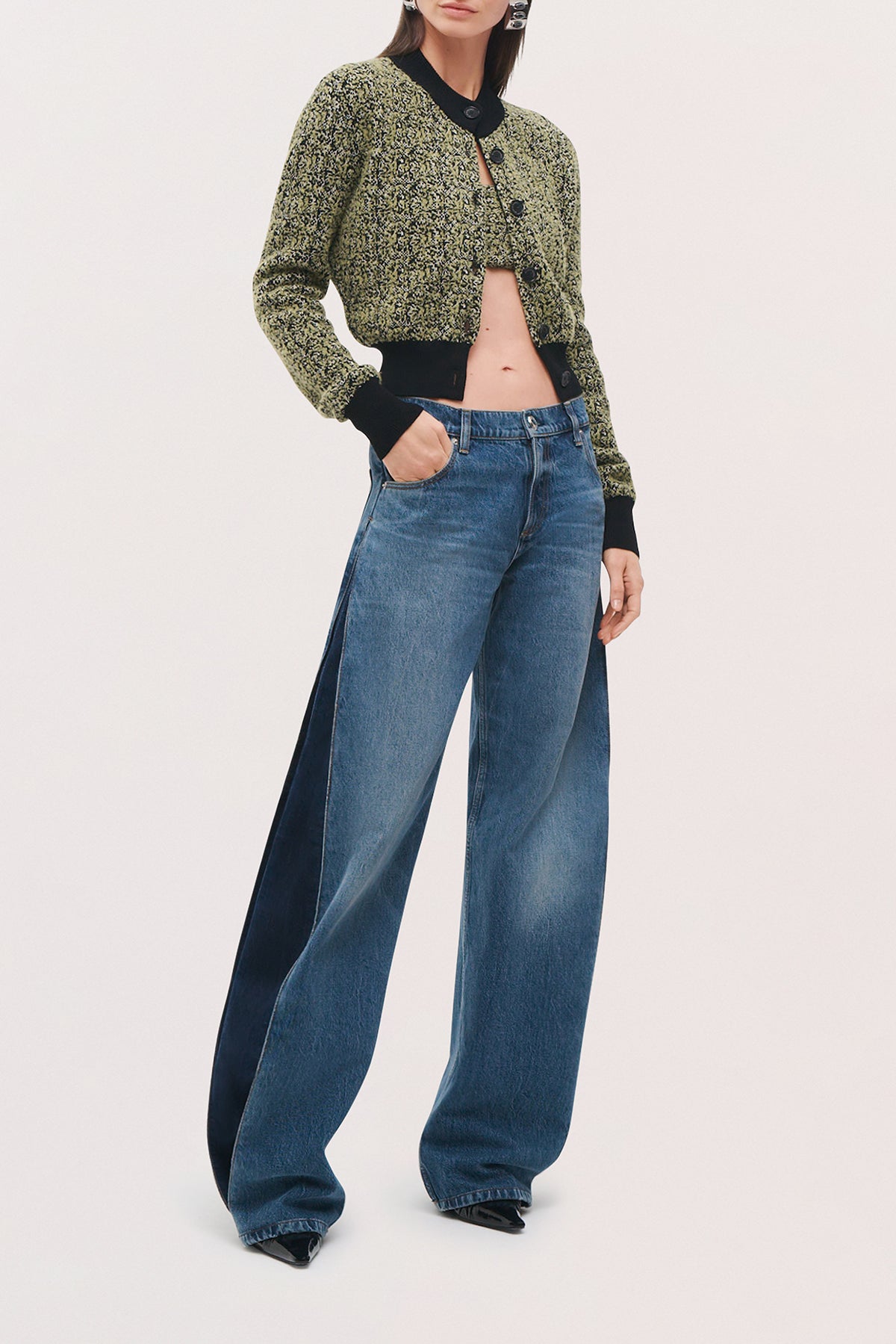 A model wearing a cropped, textured green and black cardigan with black trim, paired with wide-leg blue jeans featuring dark navy side panels, set against a plain light background.