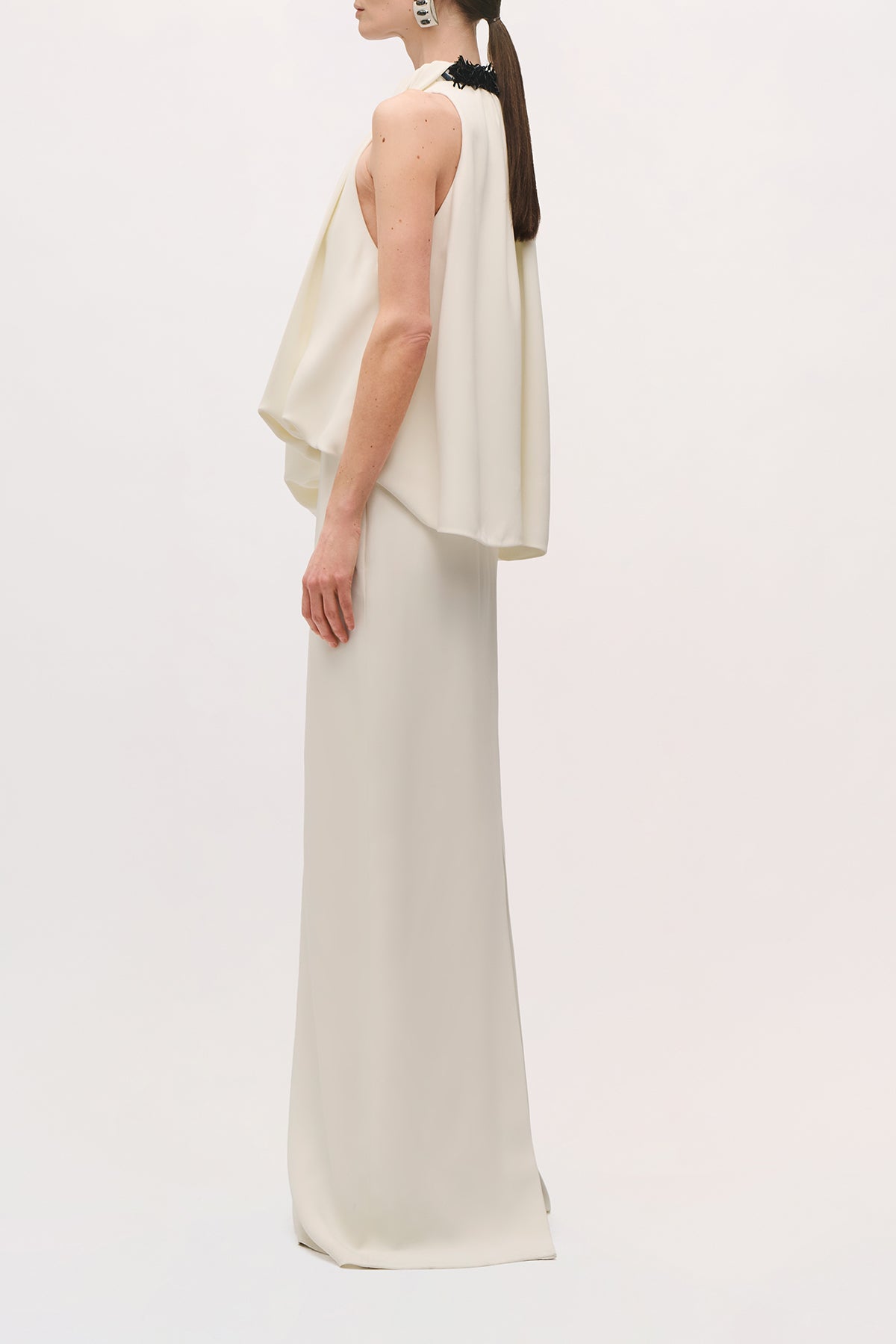Angled side view of a model wearing the Petra Gown in Ivory, showing the draped bodice, fitted waist, and straight floor-length skirt.