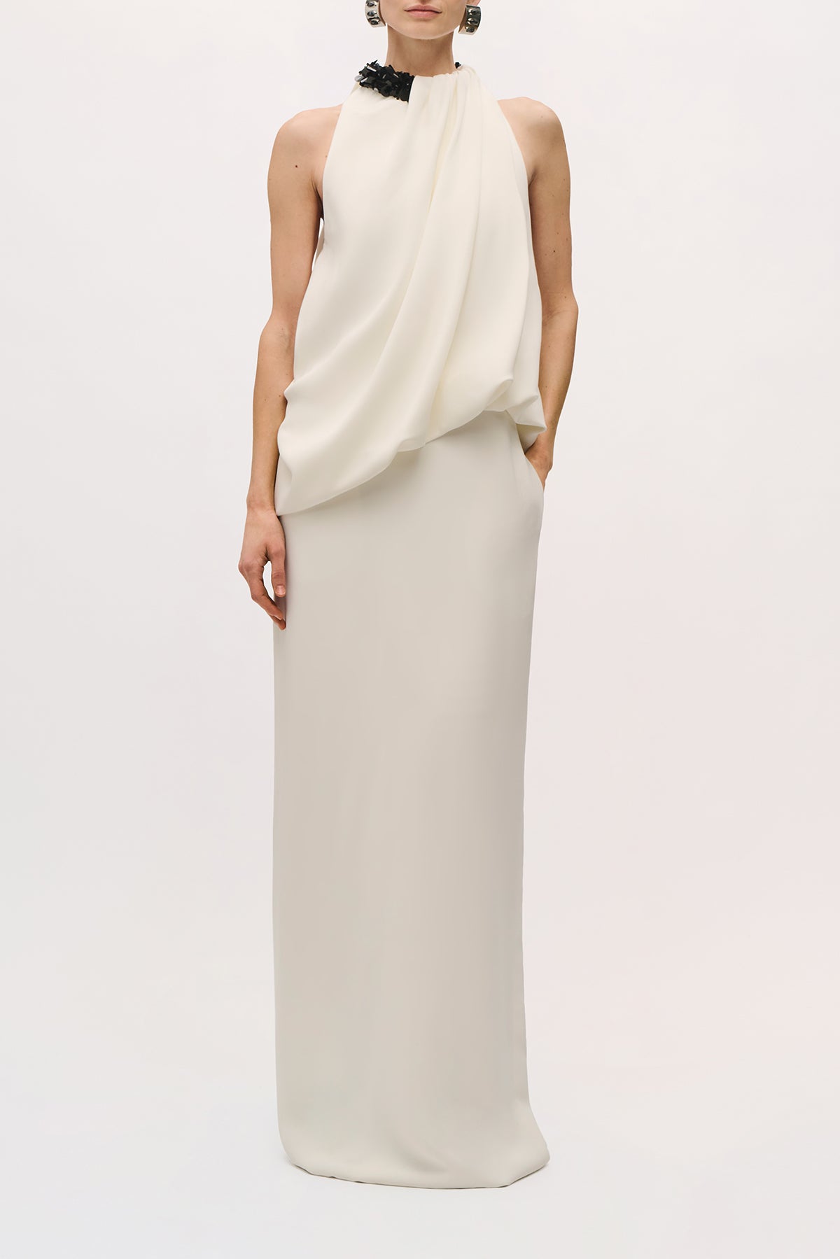 Front view of a model wearing the Petra Gown in Ivory;Black, featuring a black embellished halter neckline, draped asymmetrical bodice, and straight floor-length skirt.