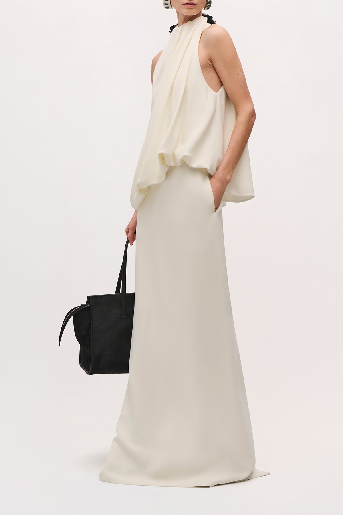 Angled side view of a model wearing the Petra Gown in Ivory;Black, and holding a black bag. Showing the draped bodice, fitted waist, straight floor-length skirt.