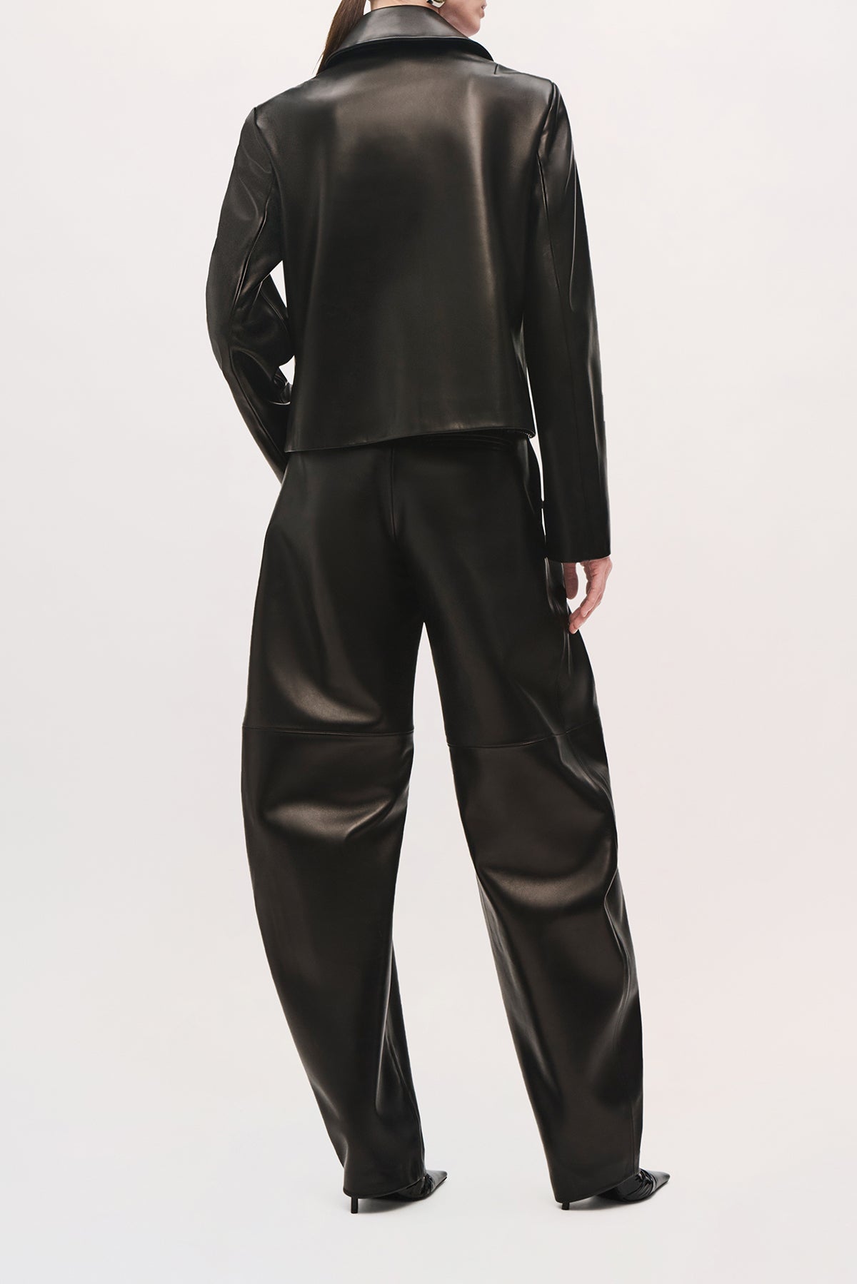 Back product view of a model wearing the Remi Jacket in black with a pointed collar, cropped fit, and one hand in the front pocket. 