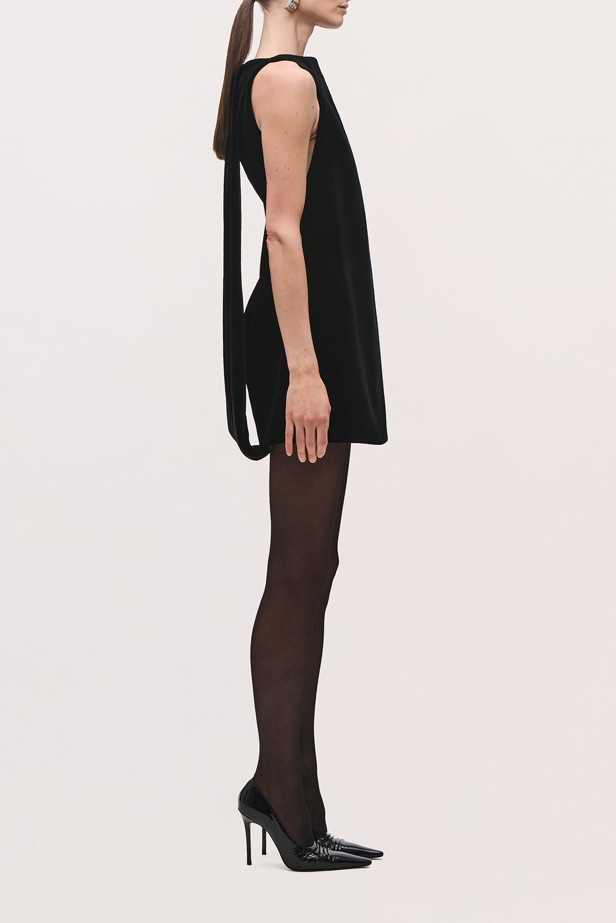 Side view of a model wearing the Sable Mini Dress in Black, a sleeveless mini dress with a straight silhouette and mantle extending down the back