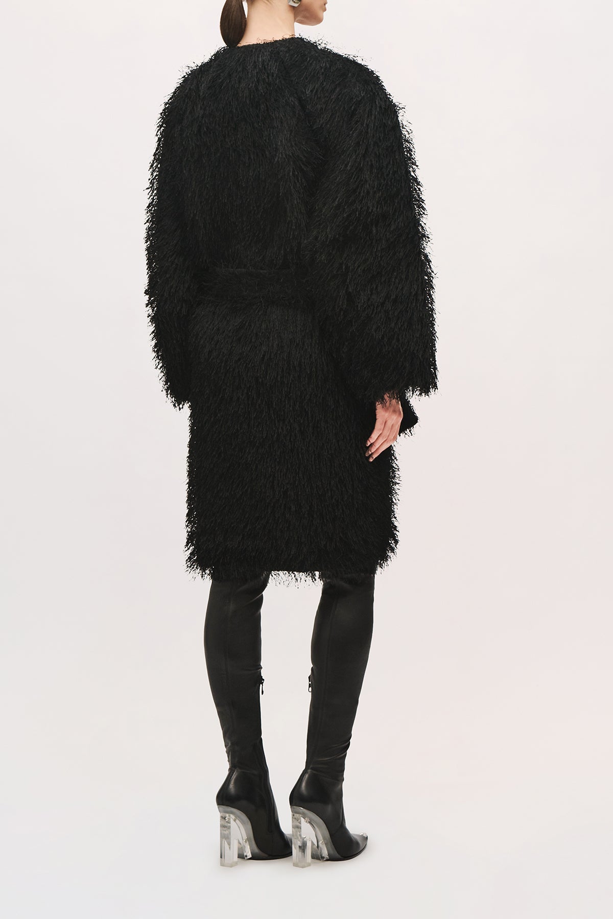 Back view of a model wearing the Sabrina Coat in Black, a knee-length coat with a textured fringe finish, relaxed cocoon silhouette, wide sleeves, and a self-tie belt at the waist.