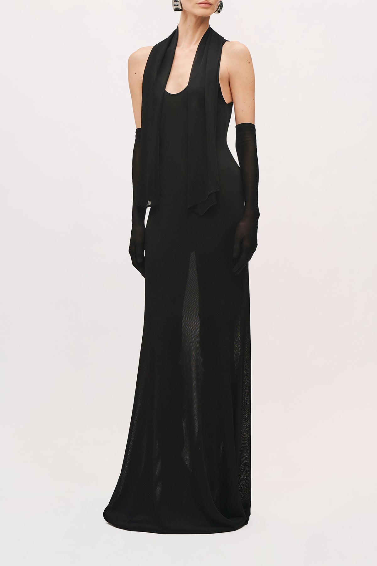 Front view of a model wearing the Siena Dress in black with a sleeveless bodice, floor-length skirt with a scoop neckline, long draped scarf panels, and a sheer textured skirt.