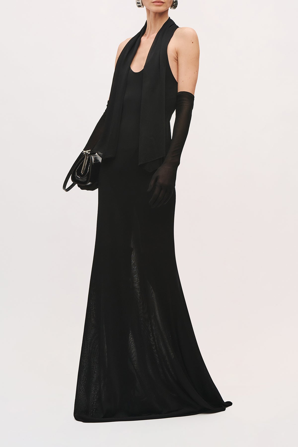 Front angle view of a model wearing the Siena Dress in Black, a floor-length black halter gown with a plunging neckline and softly draped skirt, styled with long black gloves and a black clutch. 