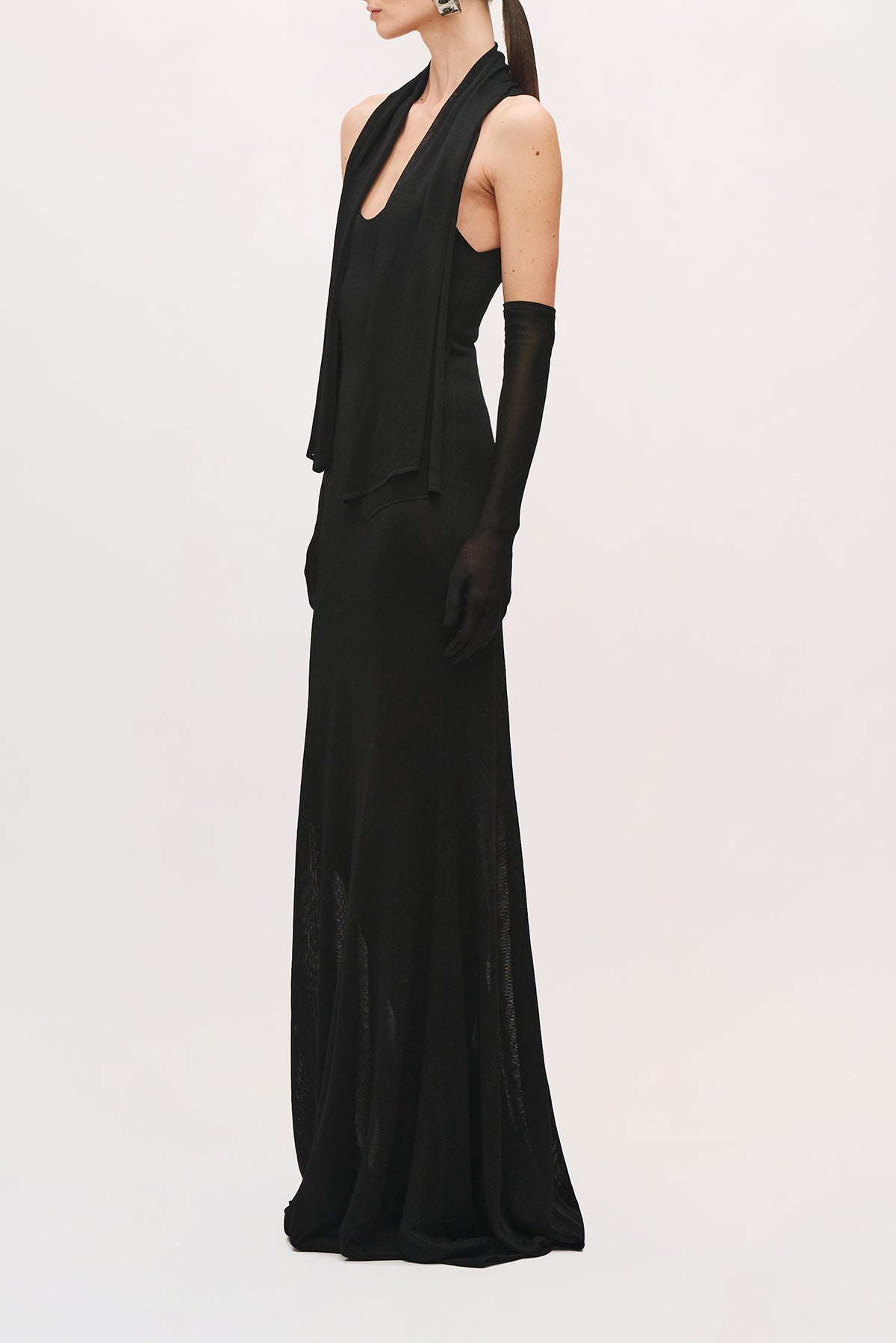 Side view of a model wearing the Siena Dress in black with a sleeveless bodice, floor-length skirt with a scoop neckline, long draped scarf panels, and a sheer textured skirt.