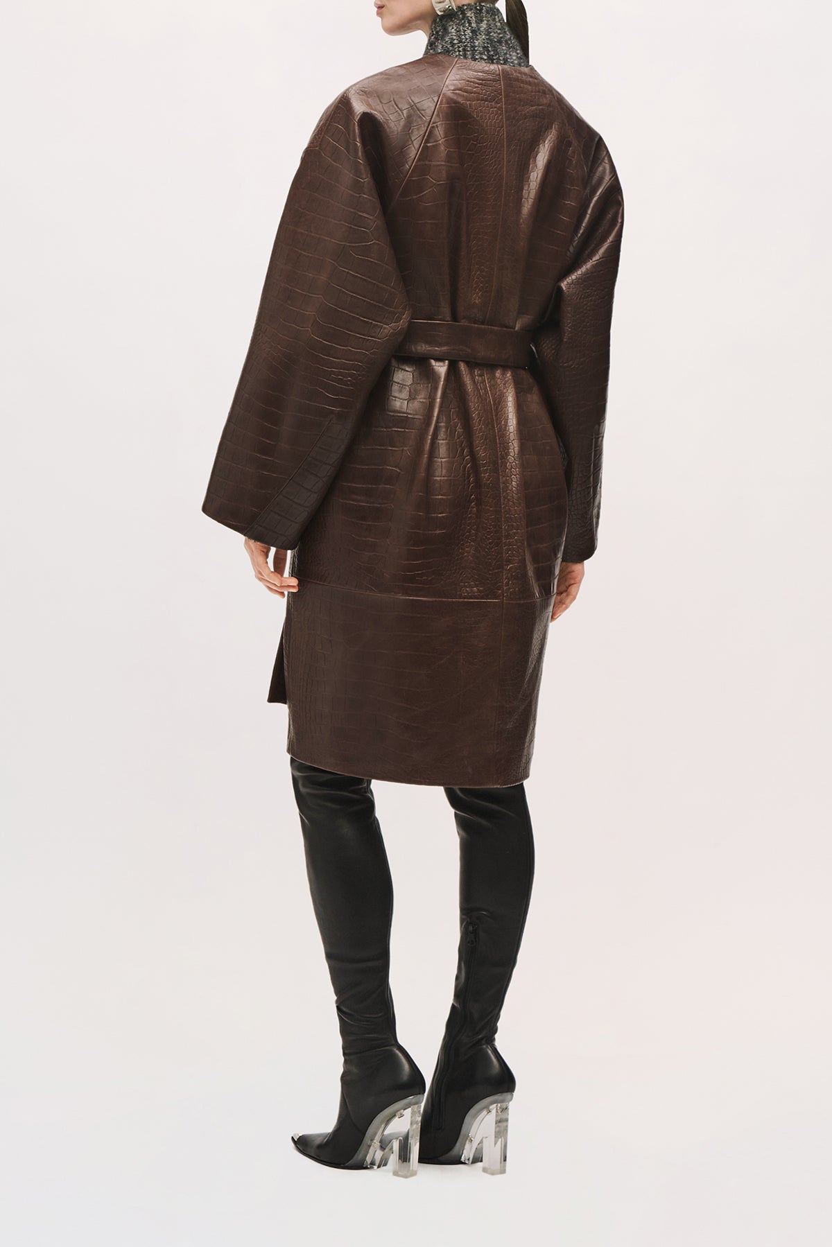 Back view of a model wearing the Trinity Coat in Brown,  a knee-length leather coat with a relaxed fit, wide sleeves, and a belted waist. 
