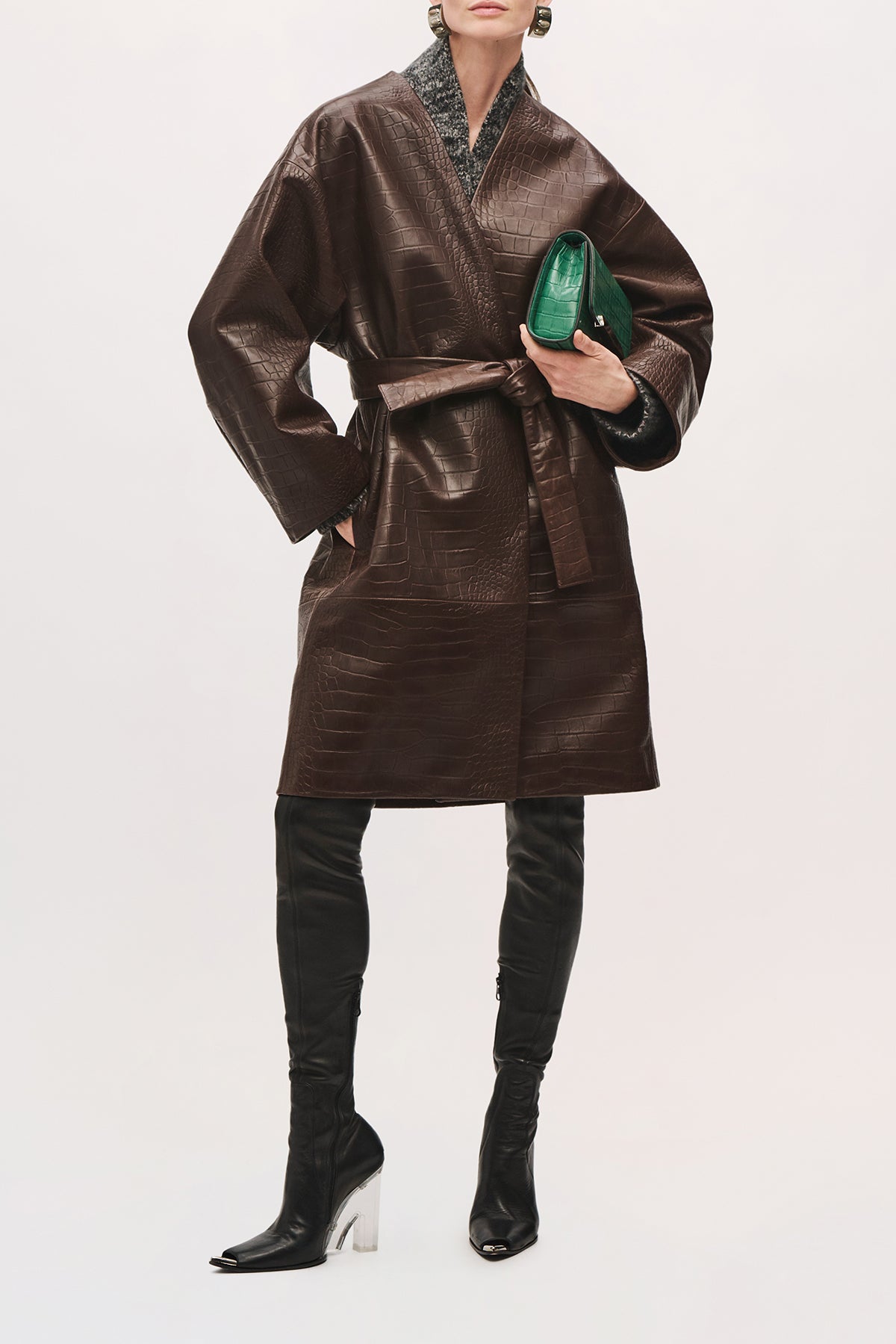 Front view of a model wearing the Trinity Coat in Brown, a knee-length leather coat with a relaxed fit, wide sleeves, and a belted waist with green accessory and purse.
