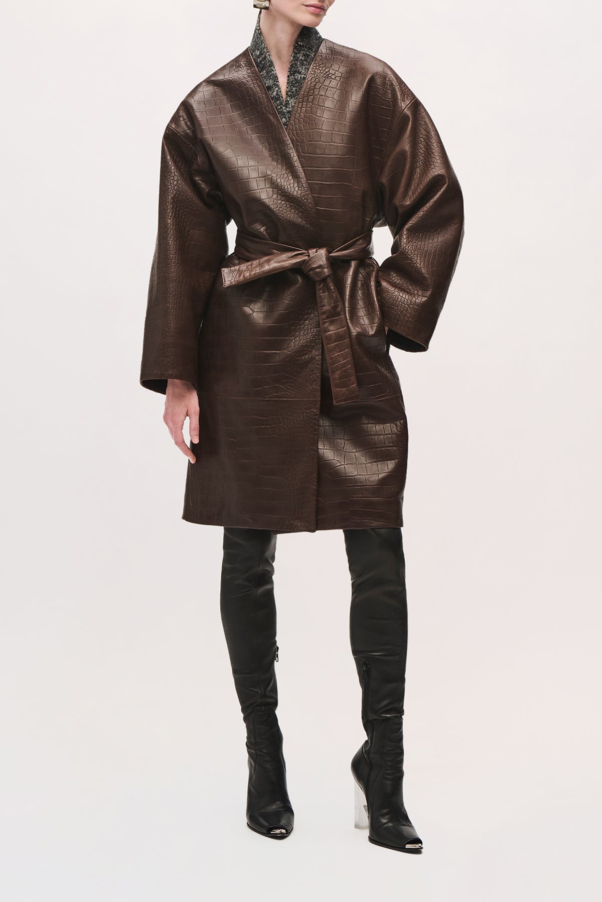 Front view of a model wearing the Trinity Coat in Brown, a knee-length leather coat with a relaxed fit, wide sleeves, and a belted waist.
